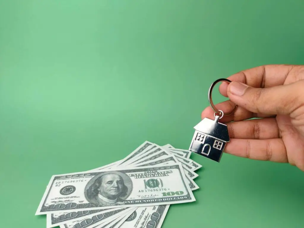 One hundred dollar bills and a person holding a keychain in the shape of a house 