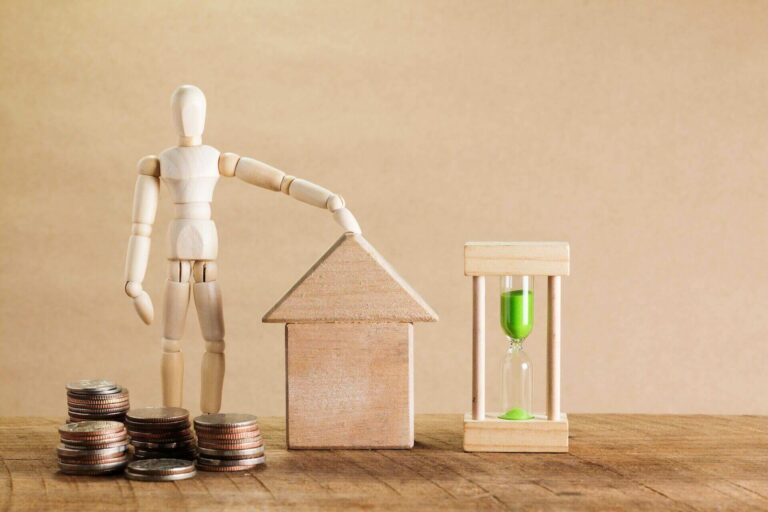 A wooden doll with a hand on a wooden home next to an hourglass and some coins